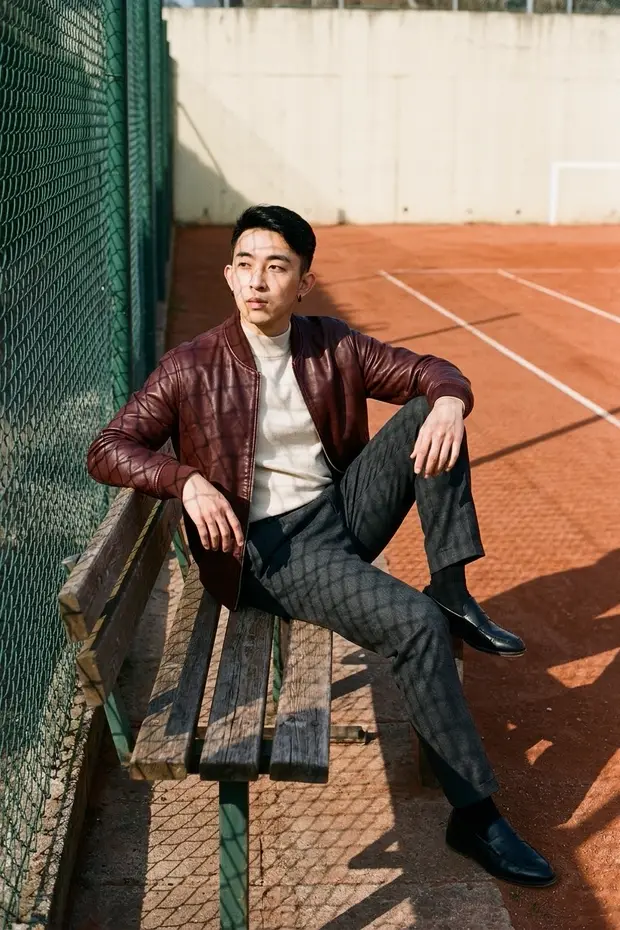 A man in a burgundy leather jacket sits on a wooden bench beside a wire fence and red tennis court.